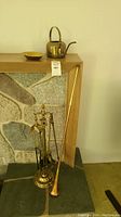 Photo showing brass teapot, brass dish, brass fireplace tool set with stand, and copper horn arranged on and near wooden mantel with stone backdrop.