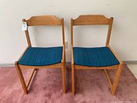 Front view of two vintage wood chairs with blue cushions on a pink carpet, showing the wood grain and design of the loose backrest.