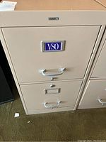 Close-up of one beige metal filing cabinet showing two drawers, handles, lock, and a blue and white VSO label on the front.