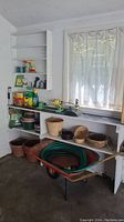 Wide shot of the gardening supplies on white shelves, with planter pots, baskets, garden sprays, and a wheelbarrow underneath.