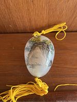 Decorative hanging egg with landscape painting and yellow tassels, photographed against wooden background.