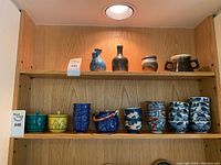 Full shelf view showing two small brown bottles, a small bowl with handle, and multiple small cups in various traditional Japanese designs on two shelves.