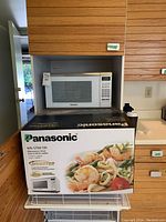 Microwave oven sitting in a kitchen cabinet with original box below it, box displays Panasonic brand and product image.
