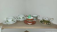 Five teacups with matching or individual saucers arranged on shelf showing variety of styles and designs.