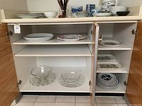 Open cabinet with multiple plates, bowls, and platters including clear glass bowls, white plates with pattern, stacked blue and white bowls.