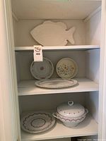 White shelf displaying the full collection of platters, bowls and plates including the fish-shaped platter on the top shelf, two decorative round plates, an oval platter, a plate with scalloped edges, and a lidded bowl on the bottom shelf.
