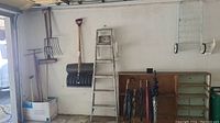 Photo shows a wider view including garden tools such as pitchfork and snow shovel hanging on wall, a metal step ladder, umbrellas in a stand, wooden shelves, and a metal drying rack on wheels.