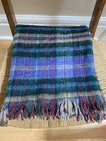 Folded wool blanket on woven chair showing plaid pattern with deep lilac, green, beige, and cinnamon colors and fringed end.