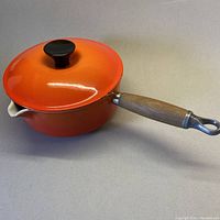 Full view of orange Le Creuset lidded pot with wooden handle and spout on a plain background.