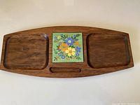 Top view of the wooden cheeseboard showing two indented trays and the colorful floral tile in the center with a small groove below it.