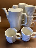 Photo showing the white ceramic tea/coffee pot with lid with several stacked and two loose large mugs with ridged detail in front.