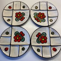 Four round ceramic plates with six divided compartments each, hand painted with red flowers and green and red dots on an off-white background, shown together