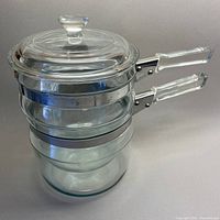 Clear Pyrex glass double boiler assembled with glass lid on top, showing stacked glass pots with stainless steel bands and clear glass handles