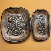 Set of two BC clay pottery trays with First Nations relief designs on warm brown background.