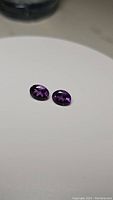 Pair of deep purple oval cut amethyst gemstones on white surface, showing color and facet detail