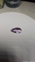 Photo of pear-shaped pink purple amethyst gemstone on a white surface showing checkerboard facets and light reflection.