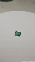 Close-up image of a loose rectangular emerald gemstone on a white surface, highlighting the emerald's color and translucency.