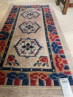 Full view of the rectangular textile area rug showing the geometric and floral sculpted patterns in red, blue, and beige colors on a cream carpet background.