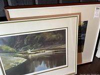 Two framed artworks leaning against dark furniture, showing landscape river scene paintings with reflective water and textured tree line, mats and frames visible