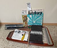 Wooden Autoharp laid flat with two green mallets on top, metal foldable music stand in background, and two instructional Autoharp books upright behind the instrument.