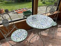 Full view of round bistro table with mosaic tile top and two chairs showing mosaic tile seats and metal scrollwork frames.