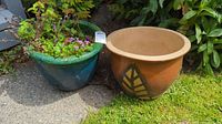 Two ceramic gardening pots side by side on outdoor ground, one green glazed with flowers and one orange-brown with leaf design, showing size and general appearance.