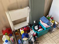 Overview image showing the white plastic step stool, ironing board, Black & Decker iron, multiple cleaning liquid bottles, and folded cleaning cloths in a green plastic container.