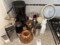 Full view of lot items on kitchen counter including coffee maker, graters, clock, bundt mold, citrus juicer, soap dispenser, cookbooks.
