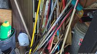 Wide view of wooden handled garden tools including rakes, axes, and brooms stored upright.