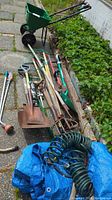 Wide view of assorted gardening tools laid on the ground including rakes, shovels, hose, and seed spreader