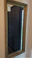 Full view of the rectangular mirror mounted on wall with gold ornate frame reflecting a nearby door and window.
