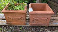 Two square terracotta pots with grapevine design, one pot containing soil and plants, the other empty, shown outdoors on wooden surface.