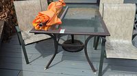 Full view of the patio table with glass top, three chairs around it, and orange umbrella resting on the table. Visible pollen on glass surface.