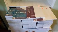 Stack of multiple unopened packages of 8.5x11 inch white resume paper made from 100% cotton, arranged in rows on a shelf.