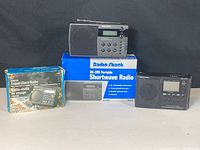Photo showing three radio clocks with two in front and one on its box in the back, with visible original packaging boxes.