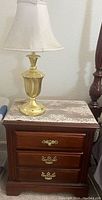 Side view of a dark wood bedside table with three drawers and brass handles, topped with a brass lamp and lace doily.