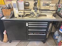 Front view of black and grey metal tool bench with four drawers and side cabinet, lathe on top (not included)