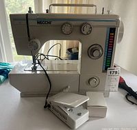 Necchi sewing machine with foot pedal and small accessory on table by window.