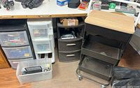 Wide view of the rolling cart with wooden top and mesh trays beside plastic storage bins stacked with miscellaneous office supplies.