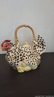 Front angle view of the ceramic chicken teapot with wicker handle and chicks around base.
