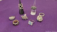 Wide shot showing all 8 assorted costume rings on purple fabric background with two rings mounted on display cones, vivid details of stones and ring designs.