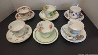 Photo showing six cups and saucers with various floral patterns and colors arranged on plain background.
