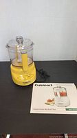 Yellow Cuisinart Mini Prep food processor with clear plastic lid and white blade, power cord attached, with instruction manual displayed on black table