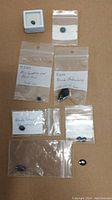 Overview of the lot showing seven loose gemstones including blue sapphire and various black stones in plastic bags and small display case.