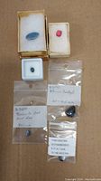 Lot overview showing multiple gemstones in boxes and plastic bags labeled with identification and sizes.
