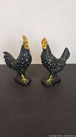 Pair of black ceramic rooster figurines with white speckles and yellow detailing on feet and comb, standing on a flat base