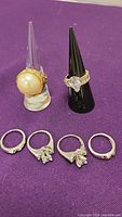 Clear display cone holds 18K gold filled faux pearl ring; black cone holds gold filled heart-cut stone ring; four white gold rings arranged below.
