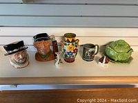 Front view of all items on a counter including Toby Jugs, Denby mug, floral vase, small house figurine, and cabbage ceramic set.