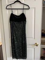 Full front view of the cocktail dress hanging on a hanger, showing black velvet top with thin spaghetti straps and green sequined skirt