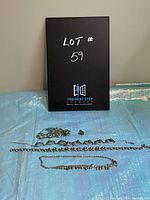 View of entire lot showing lily pattern necklace, bracelet, earrings, and two additional silver-colored necklaces laid out horizontally on blue fabric background with black lot board behind